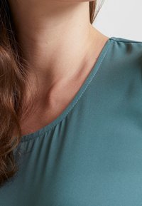 Blouse turquoise à encolure dégagée, en tissu doux et lisse avec un léger froncé au niveau de la couture. Le design est simple et élégant.
