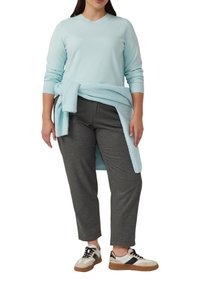 Persoon in een lichtblauwe langemouwen top met een bijpassende trui om de taille geknoopt, donkergrijze broek en witte sneakers met zwarte strepen.