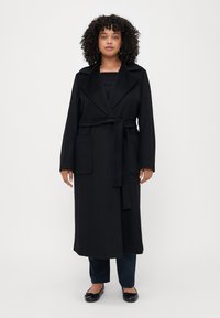 Cappotto lungo nero in morbido tessuto, con colletto a scialle, cintura da annodare e due tasche applicate. Il modello indossa una maglietta nera, pantaloni scuri e ballerine.
