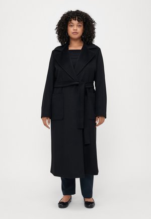 Black long coat in soft fabric, featuring a shawl collar, tie belt, and two patch pockets. Model wears black top, dark pants, and flats.