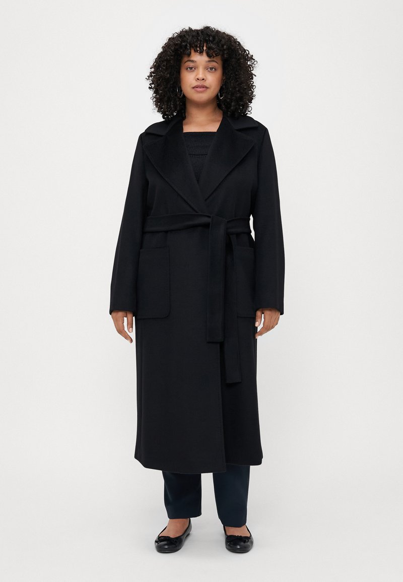 Cappotto lungo nero in morbido tessuto, con colletto a scialle, cintura da annodare e due tasche applicate. Il modello indossa una maglietta nera, pantaloni scuri e ballerine.