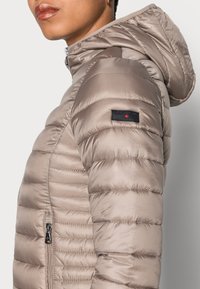 Kurtka puchowa w jasnym beżowym kolorze z satynowego materiału, z poziomym pikowaniem, kieszeniami na zamek i kapturem. Ma czarną metkę z czerwonymi akcentami na rękawie.