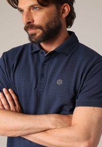 Polo en bleu marine avec un motif texturé, patte de boutonnage à trois boutons et col et poignets côtelés. Présente un logo brodé sur la poitrine.