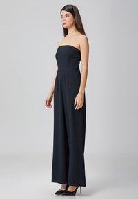 Jumpsuit noir, sans bretelles, avec un corsage ajusté et des pantalons évasés, fabriqué à partir d'un tissu lisse. Assorti à des talons aiguilles noirs.