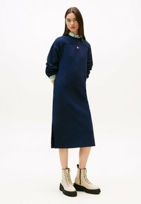 Marineblaue Langarmkleid mit Rundhalsausschnitt und seitlichen Schlitzen, kombiniert mit beigen Schnürstiefeln im Combat-Stil mit derben schwarzen Sohlen.