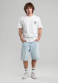 Camiseta de algodón blanca con gráfico en el pecho izquierdo, pantalones cortos de mezclilla azul claro y zapatillas blancas con calcetines de acento verde.