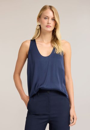 Blond vrouw met grote gouden oorbellen, marineblauwe mouwloze satijnen top en bijpassende marineblauwe broek tegen een effen achtergrond.