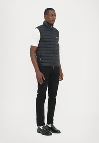 Schwarze Steppweste mit hohem Kragen, Reißverschluss und seitlichen Taschen, getragen über einem weißen Hemd, kombiniert mit schwarzen Hosen und schwarzen Slippern.