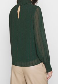 Blouse verte transparente avec rayures texturées verticales, col montant, ouverture en trou de serrure dans le dos, et manches longues avec poignets froncés.