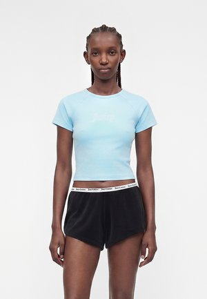 Νεαρή γυναίκα με πλεξούδες, φοράει ανοιχτό γαλάζιο εφαρμοστό crop top και μαύρα σορτς με λωρίδα μέσης με λογότυπο, στέκεται μπροστά σε απλό φόντο.