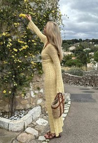 Γυναίκα με μακρύ κίτρινο φόρεμα που φτάνει για κίτρινα λουλούδια πάνω σε μια πέτρινη τοιχοποιία, κρατώντας μια υφαντή τσάντα, με ένα χωριό και συννεφιασμένο ουρανό στο φόντο.