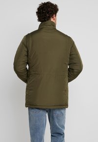 Olivgrön pufferjacka med hög krage, strukturtyg och elastiska ärmslut. Bakifrån visas en slät yta utan synliga logotyper.