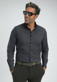 Homme portant une chemise à motifs sombres avec boutons, une ceinture tressée noire, un pantalon sombre et de grandes lunettes de soleil noires, les mains dans les poches, souriant légèrement.