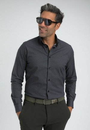 Homme portant une chemise à motifs sombres avec boutons, une ceinture tressée noire, un pantalon sombre et de grandes lunettes de soleil noires, les mains dans les poches, souriant légèrement.