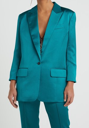 Vrouw draagt een satijnen blazer in teal en bijpassende broek, met gedeeltelijk zichtbaar metallic topje eronder en hangende oorbellen.
