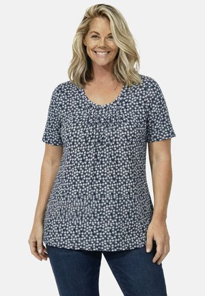 Mujer sonriente con cabello rubio hasta los hombros, lleva una blusa de manga corta azul marino con flores y jeans azul oscuro, de pie.