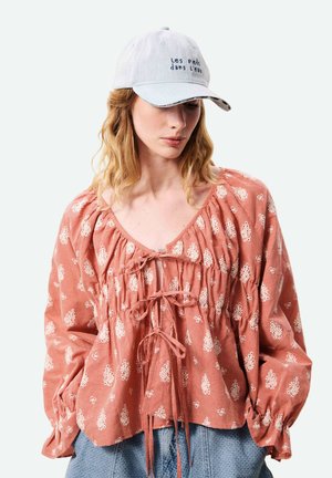 Femme portant un chemisier rose à fleurs avec des détails noués et une casquette gris clair avec un texte noir, regardant vers le bas.