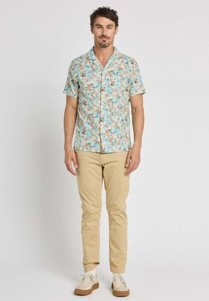 Homme debout, portant une chemise à manches courtes bleu et beige à fleurs avec boutons, un pantalon beige, des chaussettes blanches et des chaussures beiges avec semelles en caoutchouc.