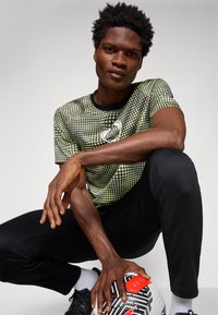 Camiseta deportiva verde y negra con estampado de puntos y cuello redondo, combinada con pantalones deportivos negros. La persona sostiene un balón de fútbol con patrón.