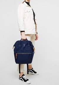 Námorně modrý batoh vyrobený z texturované látky, se hnědými koženými akcenty, zipovou přední kapsou a dvojitými ramenními popruhy.