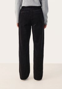 Pantalon large en velours côtelé noir avec une surface texturée, comportant deux poches arrière et une taille haute. Porté avec un pull gris.