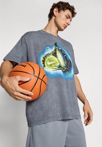 Ung mand holder en orange basketball iført en grå skjorte med et lysende troldmand- og basketballgrafik, stående mod en ensfarvet baggrund.