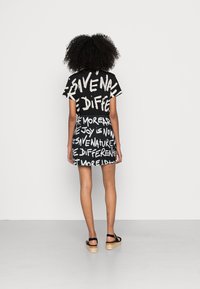 Vestido negro y crema con mangas cortas, con patrones de texto en negrita, corte ligeramente ajustado y dobladillo acampanado. Se lleva con sandalias negras.