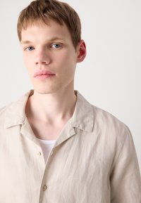 Beige button-up shirt with a soft texture, short sleeves, and a relaxed collar. Features subtle logo embroidery on the chest.