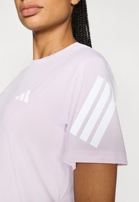 Jasnofioletowa sportowa koszulka z białymi paskami na rękawie i minimalistycznym logo na piersi. Gładka, oddychająca tkanina.