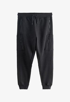 Mann trägt schwarze Jogginghosen mit seitlichen Reißverschlusstaschen und schwarze Freizeitschuhe mit beigen Sohlen und Schnürsenkeln, steht an einer beigen Wand.