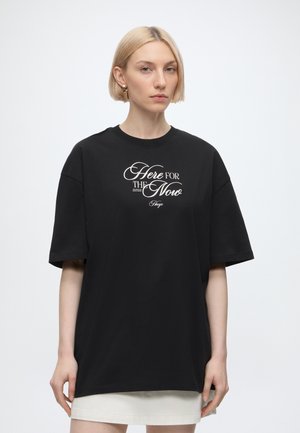Femme blonde portant un t-shirt noir oversize avec un texte blanc lisant « "Here for the Now" » et une jupe blanche, debout devant un fond uni.