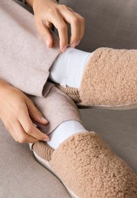 Beige, flauschige Hausschuhe mit einer weichen, texturierten Außenseite und gerippten Akzenten, getragen über weißen Socken und kombiniert mit hellrosa Freizeitabwear.
