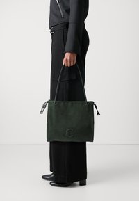 Borsa tote in suede verde con texture liscia, manici corti e dettaglio del logo. Indossata con pantaloni neri a gamba larga e stivali con tacco.
