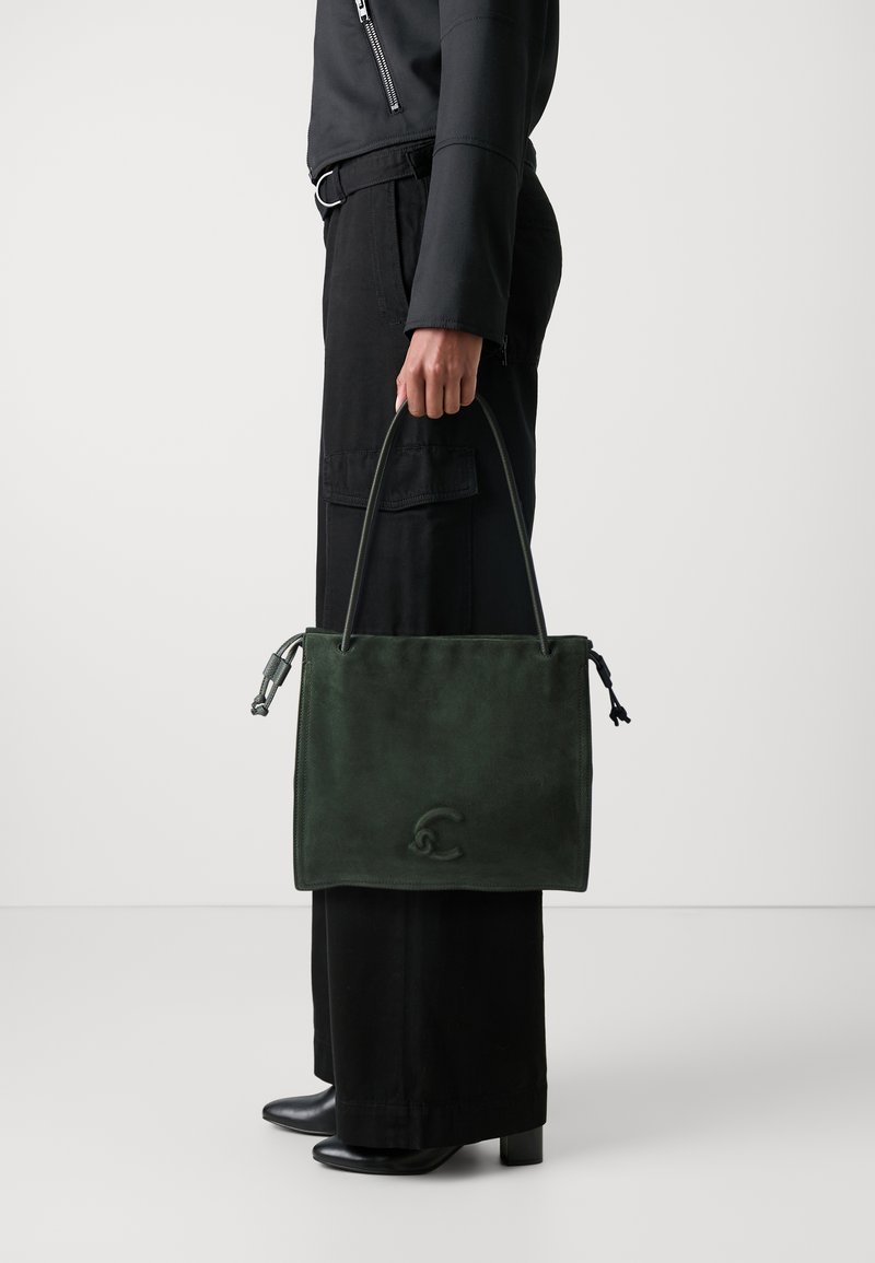 Borsa tote in suede verde con texture liscia, manici corti e dettaglio del logo. Indossata con pantaloni neri a gamba larga e stivali con tacco.