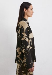 Blouse à motifs floraux noir et beige, à coupe ample, à manches longues et à col, confectionnée en tissu doux.