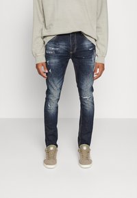 Tmavě modré džíny z denimu s opotřebovanými detaily, dobře padnoucí design a zlatými švy, zkombinované s béžovými teniskami a světlým šedým svetrem.