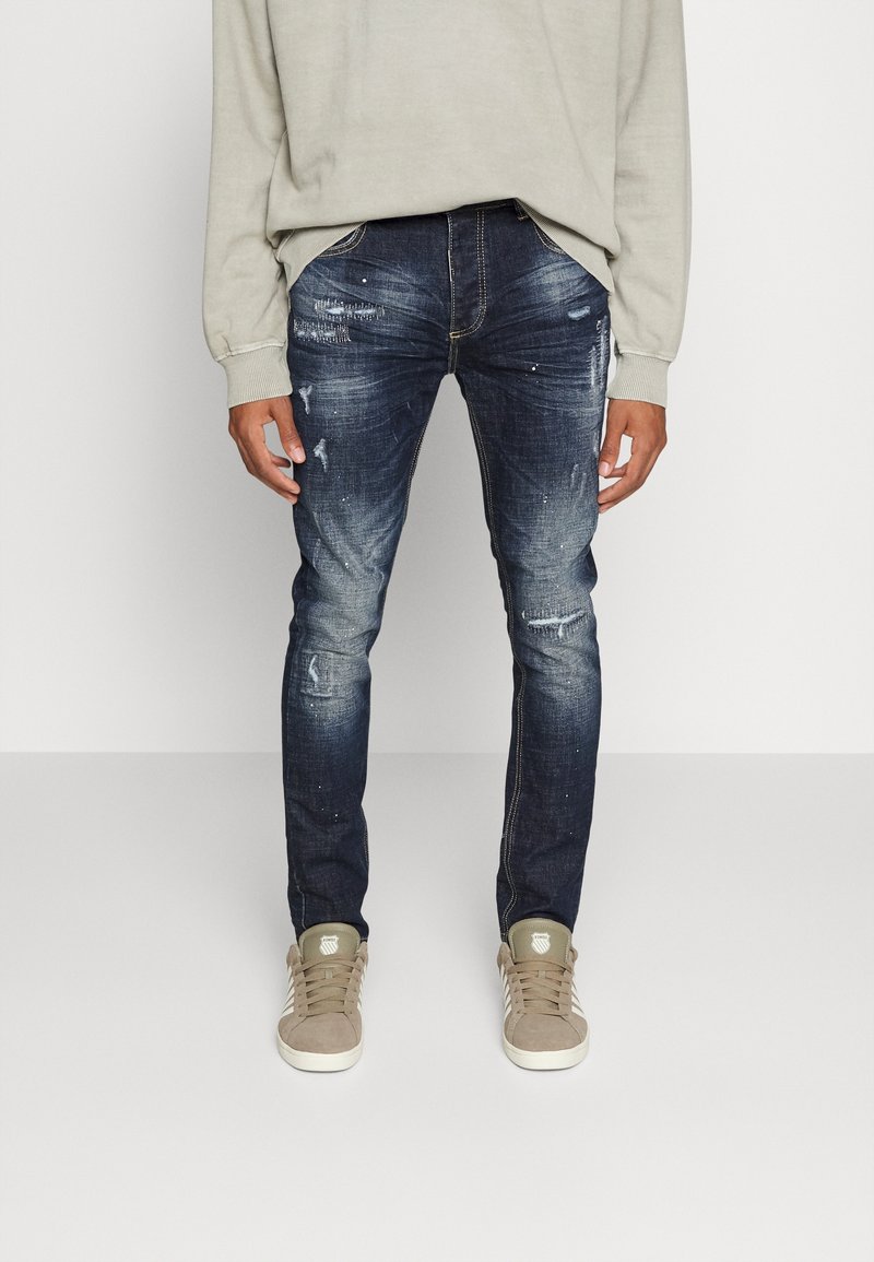 Tmavě modré džíny z denimu s opotřebovanými detaily, dobře padnoucí design a zlatými švy, zkombinované s béžovými teniskami a světlým šedým svetrem.