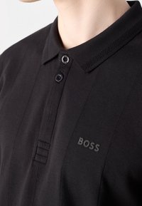 Polo noir à col avec deux boutons et logo "BOSS" en relief subtil sur la poitrine gauche, porté par une personne.