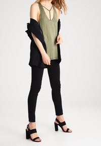 Canotta verde oliva con scollatura a V profonda e spalline incrociate, abbinata a una giacca nera, jeans skinny neri e sandali con tacco a blocco neri.