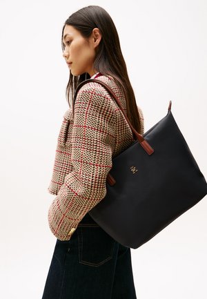 Donna con giacca a quadri marrone porta una grande borsa tote nera con manici marroni sulla spalla, su sfondo bianco.