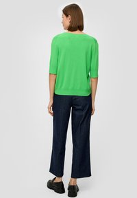 Helle grüne Sweater mit ¾-Ärmeln, kombiniert mit dunkelblauen weit geschnittenen Jeans und schwarzen Schuhen, von hinten betrachtet.
