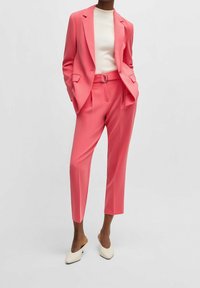 Růžový přesně střižený oblek se sakem na jedno knoflíkové zapínání, kalhotami rovného střihu a bílým žebrovaným topem. Obsahuje pásek se stříbrnou sponou a špičatou obuv.