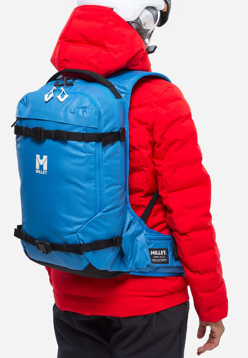 Blauer Hirse-Rucksack mit schwarzen Akzenten, verstellbaren Trägern, vorderen Schnallen und Logo; getragen über einer roten Daunenjacke auf einer Person.