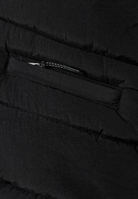 Svart pufferjacka med detaljer kring en zippered ficka. Materialet verkar mjukt med en något texturerad yta. Sytt accent synligt längs sömmarna.