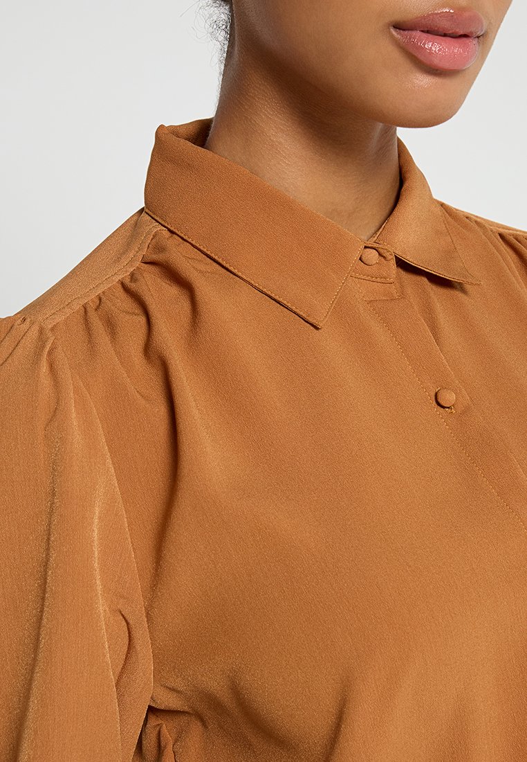 Bruine knoop-blouse met een gestructureerde kraag, zachte textuur en pofmouwdetails. Bevat kleine knopen aan de voorkant voor sluiting.