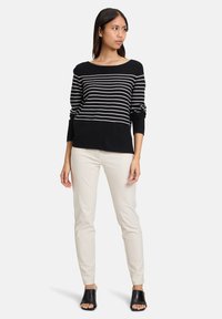 Gestreifter schwarz-weißer Pullover mit Bootsausschnitt, langen Ärmeln und einem einfarbigen schwarzen Bund am unteren Ende, kombiniert mit cremefarbenen, figurbetonten Hosen und schwarzen Mules.