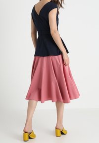 Mujer vestida con un top negro ajustado de espalda en V, una falda midi rosa acampanada y mules amarillos de tacón bloque, de pie contra un fondo blanco liso.
