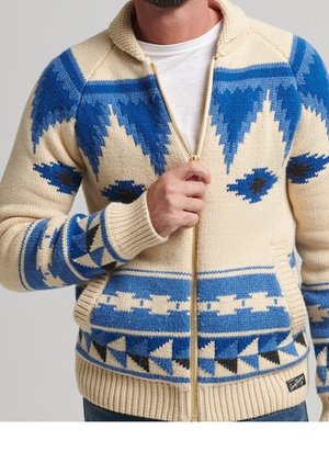 Cardigan con zip di colore crema, caratterizzato da motivi geometrici blu e dettagli. Tessuto lavorato a maglia con polsini e orlo a costine, due tasche laterali.
