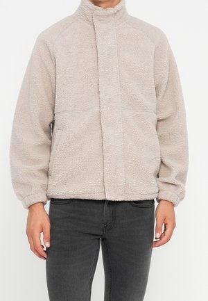 Chaqueta de forro polar beige con cuello alto y puños elásticos, llevada por una persona con vaqueros negros, de pie contra un fondo blanco.