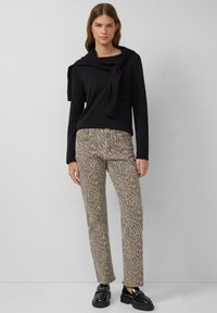 Top nero a maniche lunghe con dettagli di collo annodato, abbinato a pantaloni beige con stampa leopardata e scarpe nere in vernice, in piedi davanti a uno sfondo neutro.